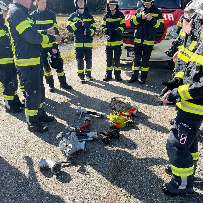 Eine Gruppe von Feuerwehrleuten in Uniformen mit reflektierenden Streifen steht im Kreis mit einigen Ausrüstungsgegenständen auf dem Boden. Ein roter Feuerwehrwagen ist in der Nähe geparkt.
