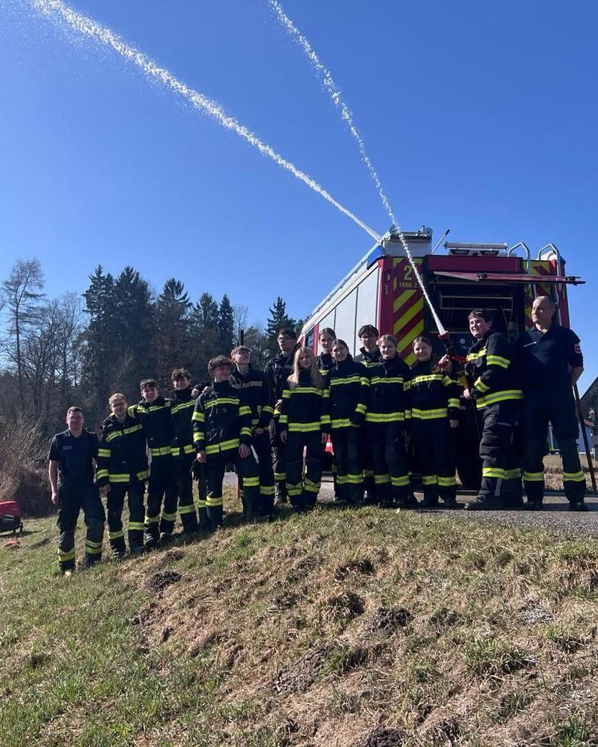Eine Gruppe von Feuerwehrleuten und jungen Personen steht draußen zusammen, wobei ein Feuerwehrauto im Hintergrund Wasser sprüht, unter einem klaren blauen Himmel.