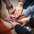 Eine Gruppe von Personen mit ihren Händen, die in einer Einheitsgeste aufeinandergestapelt sind. Sie tragen verschiedene Accessoires wie Armbänder und Ringe.