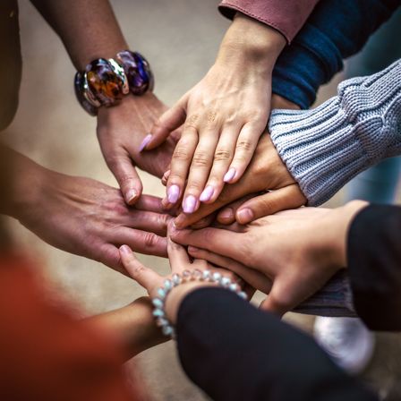 Eine Gruppe von Personen mit ihren Händen, die in einer Einheitsgeste aufeinandergestapelt sind. Sie tragen verschiedene Accessoires wie Armbänder und Ringe.