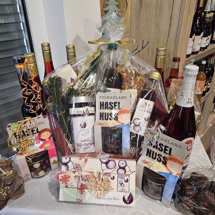 A gift basket with various products including wine bottles, candies, and other items. The basket is wrapped in plastic and has a ribbon. The products are displayed on a table in a shop.