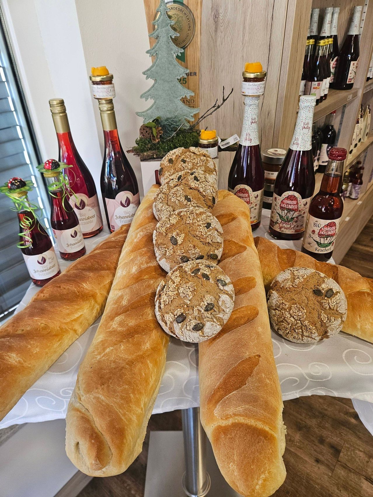 Ein Tisch mit einer weißen Tischdecke zeigt mehrere Brote, einige mit Samen, und Flaschen Wein und Sirup auf einem hölzernen Regal im Hintergrund.