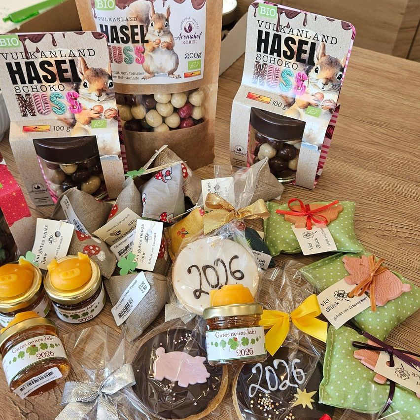 Auf einem Holztisch sind verschiedene Lebensmittel ausgestellt. Dazu gehören verpackte Pralinen, Kekse und Gläser mit Marmelade, alle mit einem 2026-Etikett. Es gibt auch Geschenktüten und eine Schachtel mit einem Eichhörnchen-Bild.
