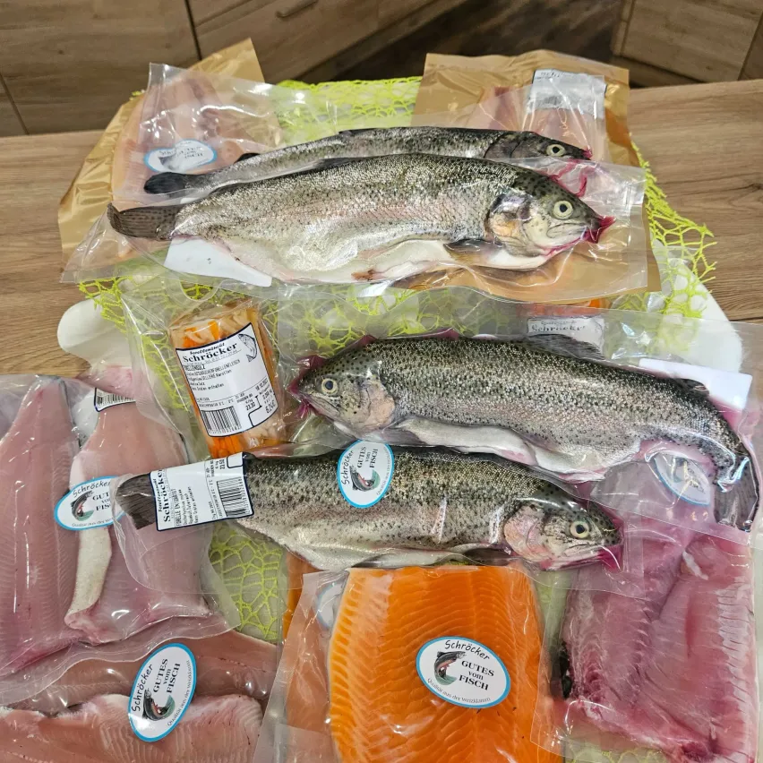 Verschiedene frische Fischprodukte, einschließlich Filets und ganze Fische, sind ordentlich in Plastiktüten auf einem Holztisch angeordnet. Jeder Fisch hat ein blaues Etikett mit weißem Text.