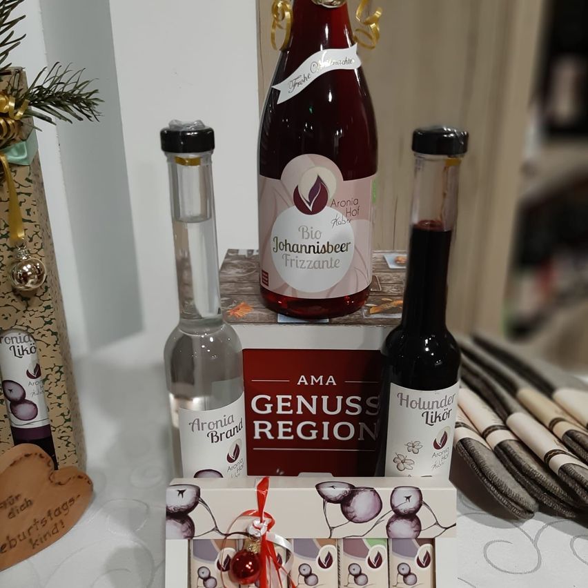 Ein Geschenkset besteht aus einer Flasche Johannisbeer Frizzante, einer Flasche Aronia Brand und einer Flasche Holunder Likör. Das Set ist mit Feiertagsschmuck und einem Herz dekoriert.