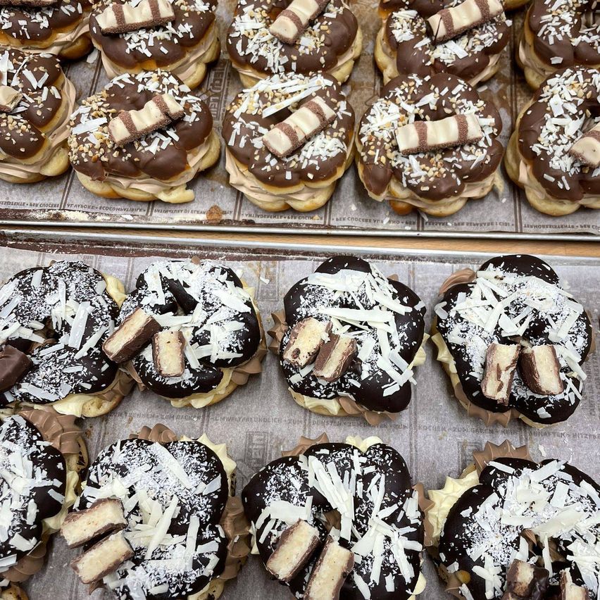 A tray of chocolate-covered donuts with Snickers bars and coconut flakes on top, arranged in two rows. Another tray below has donuts with chocolate and white flakes.