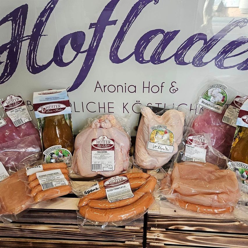 A display of various meats, sausages, and products in plastic packaging. A sign with purple text reads 'Aronis Hof & Lader'. The products include chicken, sausages, and other meats.