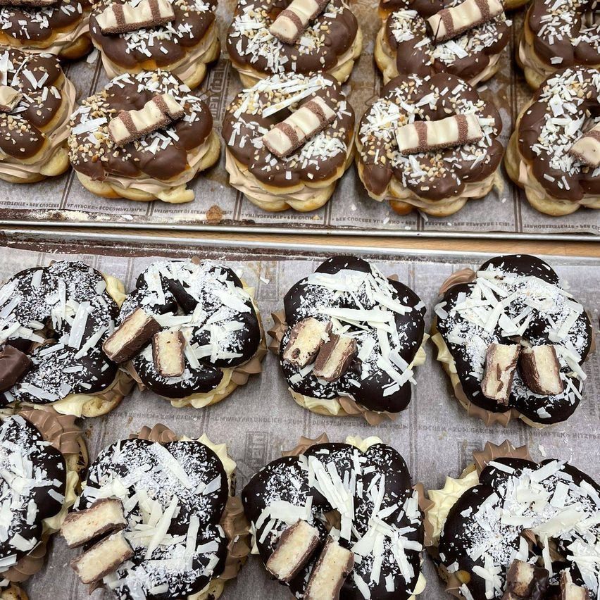 Two trays of donuts, each covered in chocolate icing and topped with shredded coconut and Kit Kat pieces. The donuts have a light, fluffy texture.