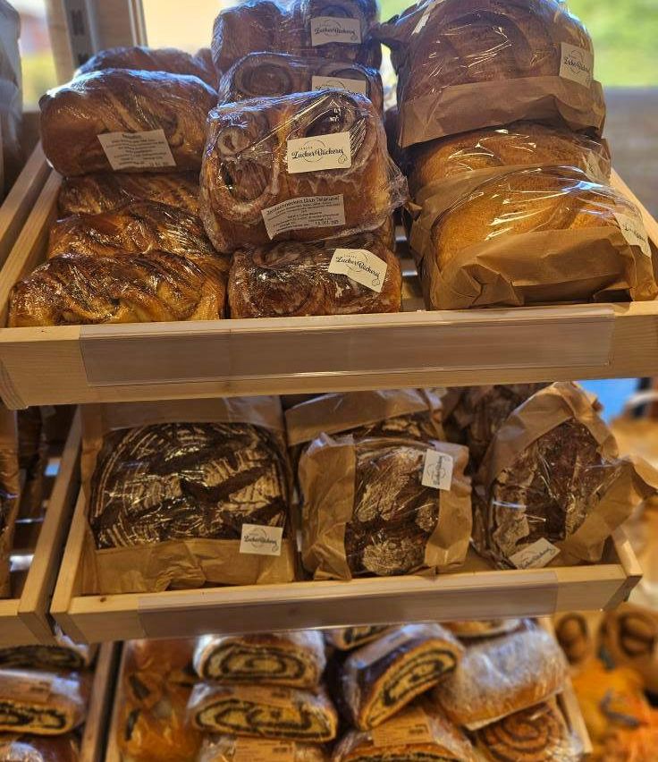 A wooden shelf displays various breads. The top layer features wrapped loaves in plastic, including some with visible labels. Below, there are more loaves in brown paper, some also labeled. Below these, there are additional loaves in brown paper, with some featuring black swirls.