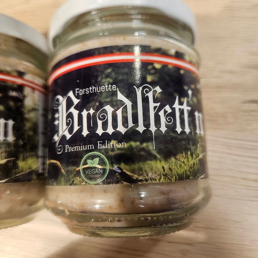Zwei Glasgläser mit weißen Deckeln und Etiketten, auf denen 'Forsthuette Bradlfett Premium Edition' steht. Das Etikett zeigt ein Bild eines Grasfeldes und ein grünes veganes Logo.