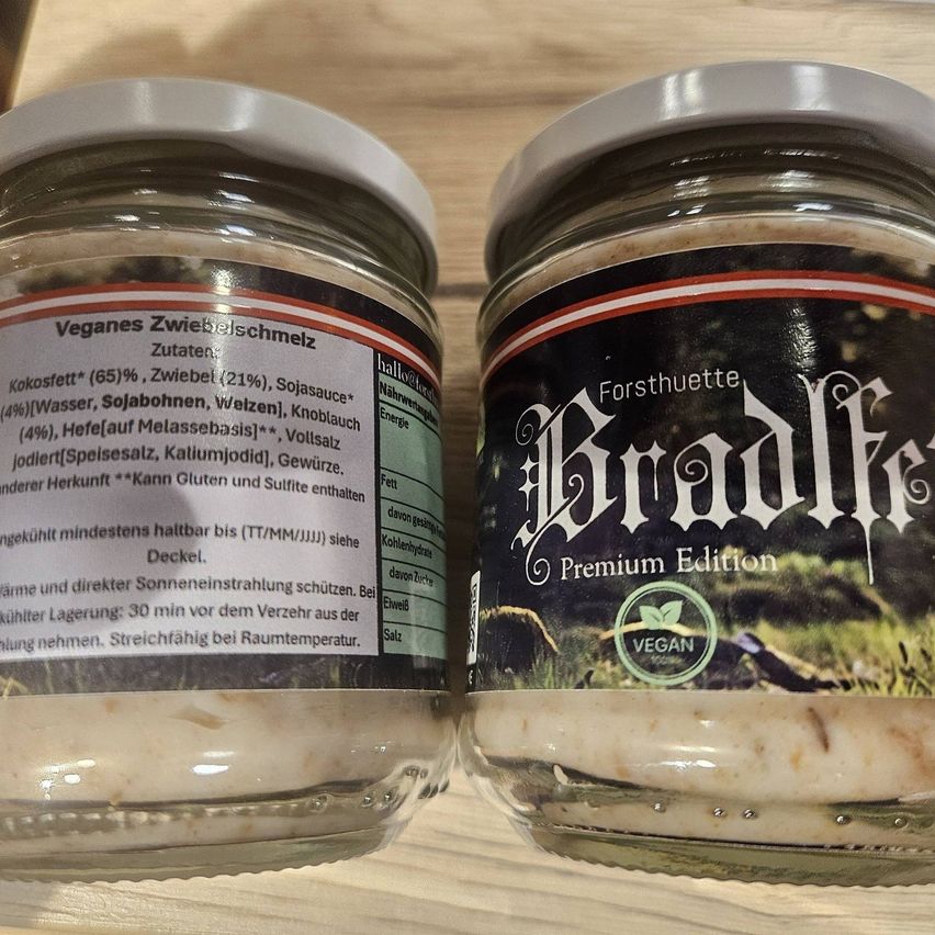 Zwei Glasgläser mit Etiketten auf einer Holzoberfläche. Das linke Glas sagt 'Veganes Zwiebelschmetz'. Das rechte Glas sagt 'Forsthuettte' und 'Brade' mit 'Premium Edition'. Beide Gläser enthalten eine weiße Substanz.