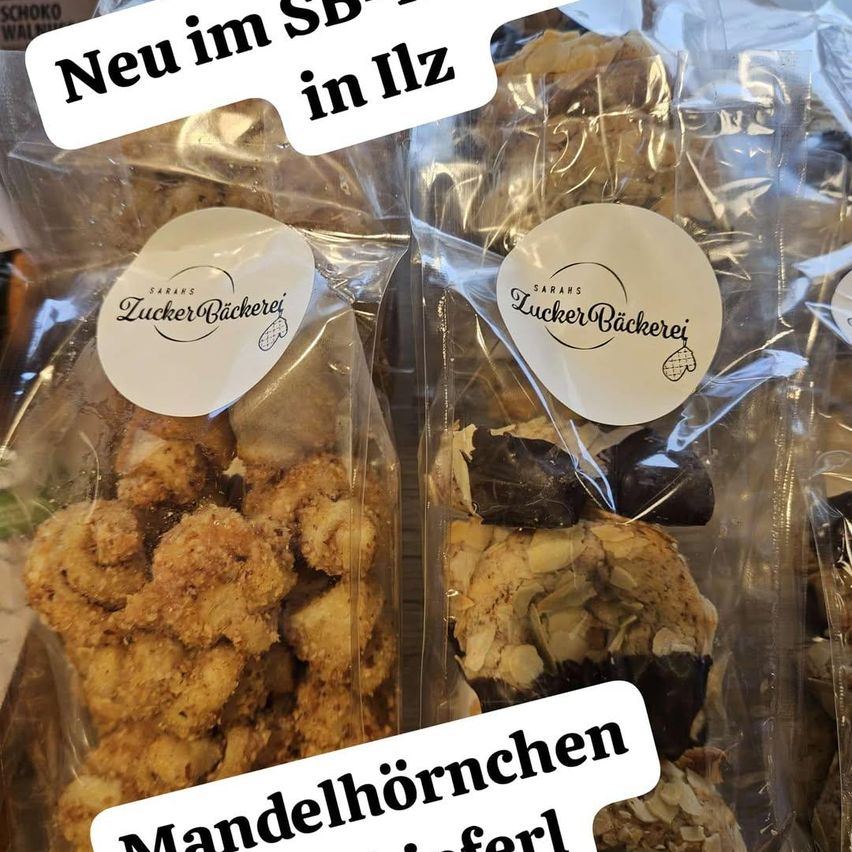 Pakete von Keksen sind abgebildet, mit Etiketten, die 'ZuckerBackerei' und 'Sarahs ZuckerBackerei' lesen. Die Produkte umfassen 'Mandelhörnchen' mit Mandeln und Schokolade. Das Bild hat einen Wasserzeichen, der 'Neu im SB in Ilz' sagt.