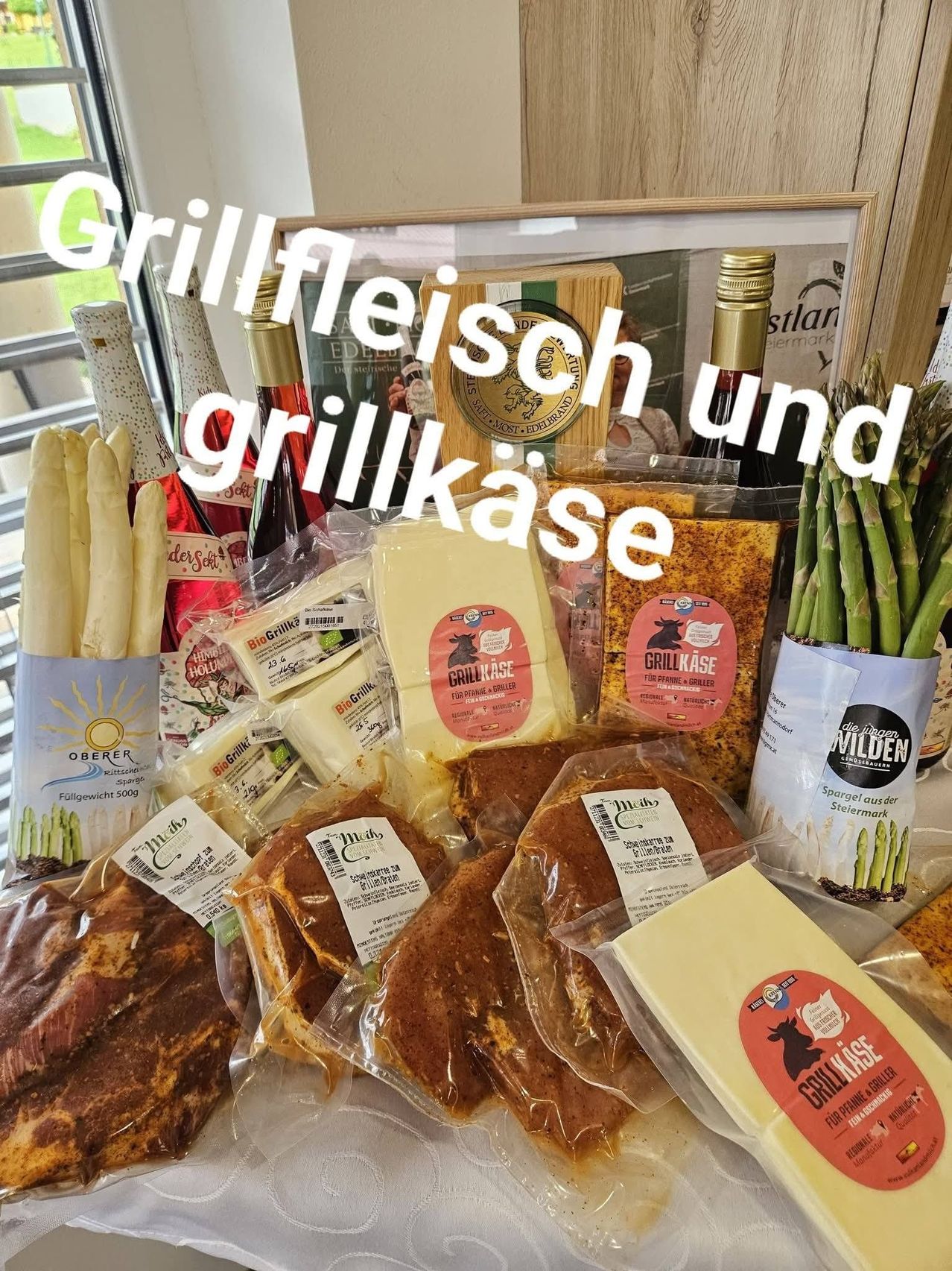 A table with various food items, including meat, asparagus, and cheese, is arranged in a visually appealing manner. The assortment includes a variety of cheese types, asparagus, and meat slices, all packaged in clear plastic. A sign with the text 'Grillfleisch und grillkase' is prominently displayed.