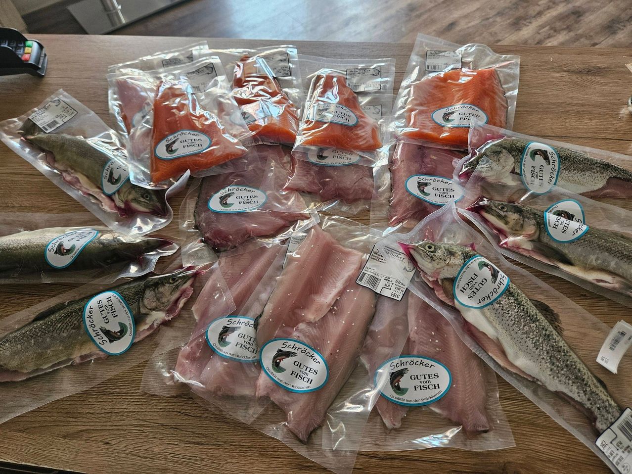 Eine Auswahl an Fischfilets, einschließlich Lachs und Forelle, ist einzeln verpackt auf einem Holztisch. Jedes Paket hat ein Etikett mit dem Text 'Gutes vom Fisch' und einem Fischlogo.