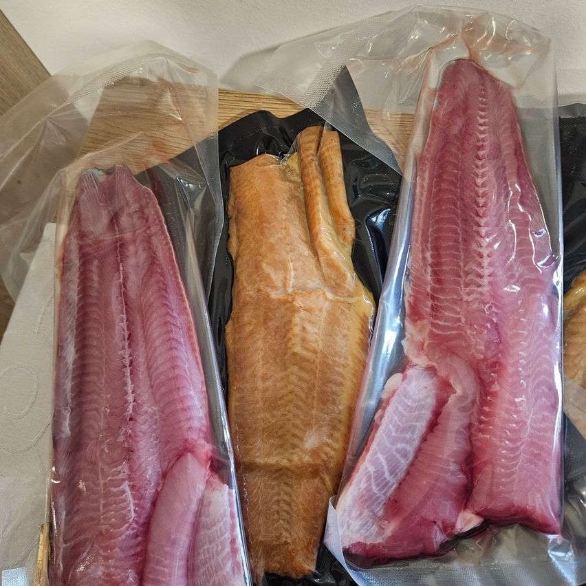 Drei Fischfilets in Plastikverpackungen, zwei rosa und eines braun, liegen auf einer Holzoberfläche. Die rosa Filets sind links, während das braune Filet in der Mitte ist.