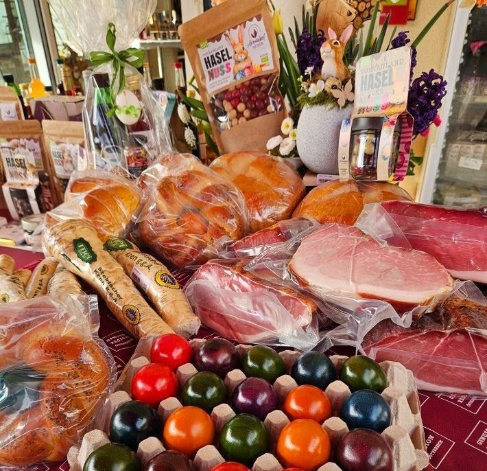 Ein Tisch voller verschiedener Lebensmittel, darunter eingewickeltes Brot, Wurstwaren und bunte Eier in einem Karton. Im Hintergrund befinden sich Blumensträuße und Werbeartikel.