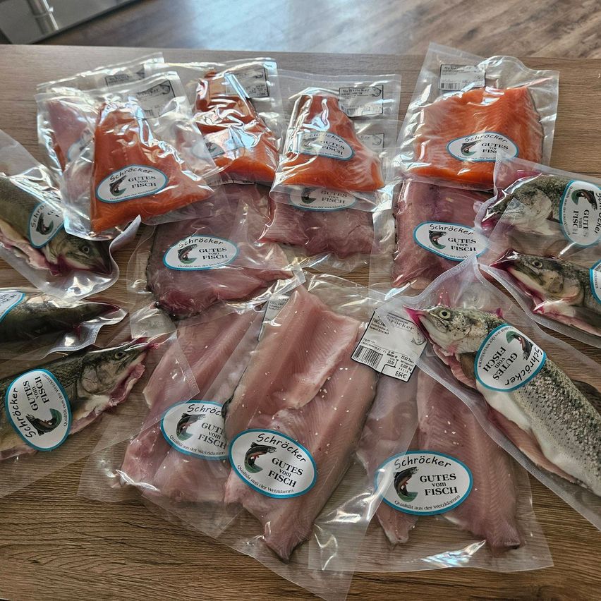 Auf einem Holztisch sind mehrere Packungen mit Fischfilets und Lachs zu sehen, alle in Plastikverpackung, jede mit einem Aufkleber, der 'Schröcker Guter Fisch' liest. Einige Packungen haben Fischköpfe, andere Fischschwänze. Der Lachs ist orange und die Fischfilets weiß.