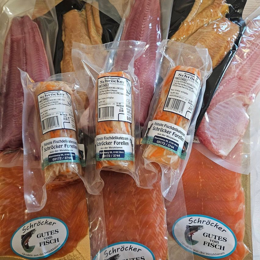 Mehrere Packungen Fisch, darunter Lachs und Forelle, sind in Plastikverpackungen. Jede Packung hat ein Etikett mit einem blauen Fischsymbol und dem Text 'Schröcker Gutes vom Fisch'. Die Fischpackungen haben unterschiedliche Größen und befinden sich auf einem Tisch.
