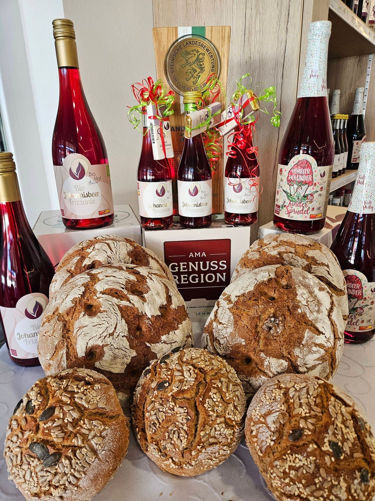 A table displays several loaves of bread with seeds on top, along with bottles of Johannisbeer Frizzante wine, some with festive ribbons. The background shows a wooden shelf with more wine bottles.