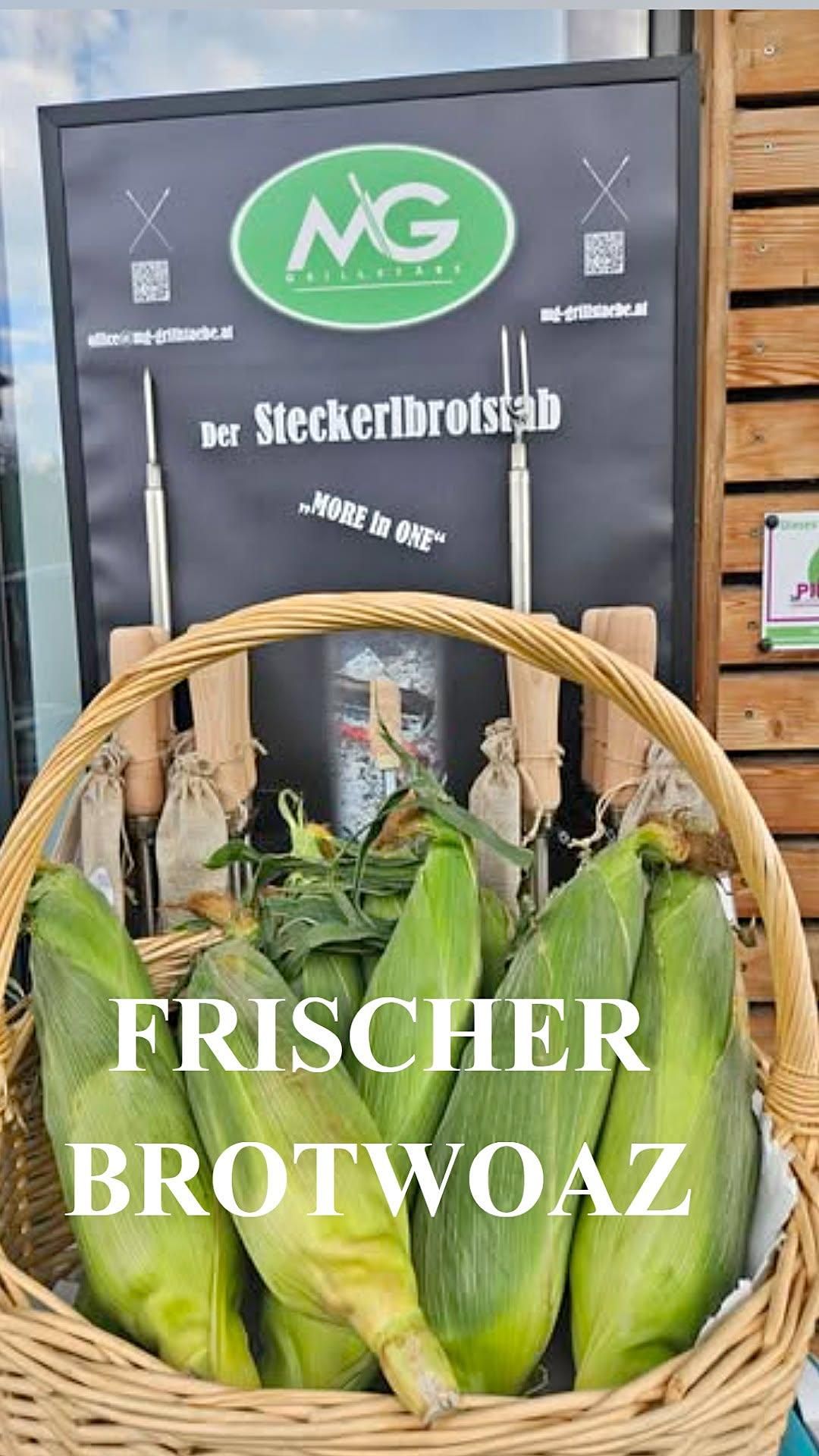 A basket of fresh corn on the cob with husks still attached, displayed in front of a sign that reads 'Der Steckerlbroststab'. The basket is placed against a wooden wall.