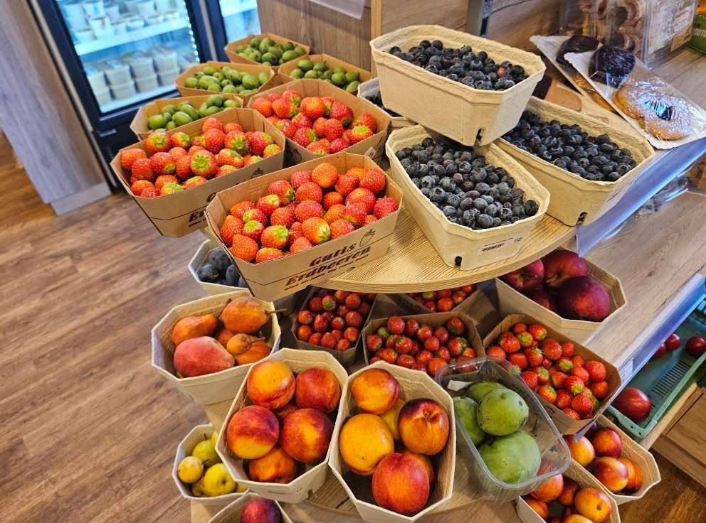 A wooden table displays a variety of fruits in cardboard boxes, including strawberries, blueberries, peaches, and pears. The setting appears to be a market or a fruit stand.
