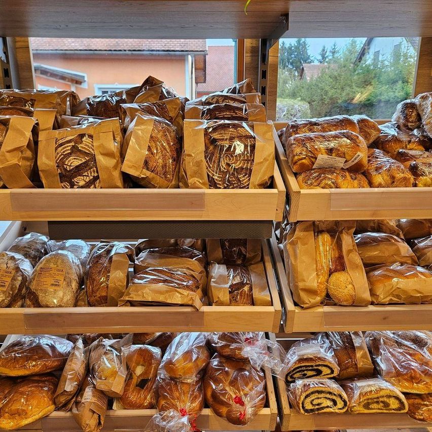 A wooden shelf displays various types of bread, including loaves wrapped in plastic and brown paper, with a view of trees and a building outside.
