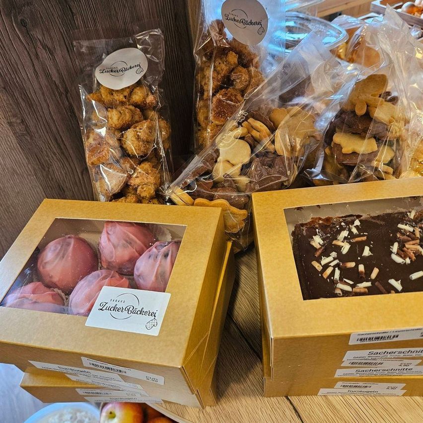 Two boxes of desserts are displayed on a wooden table. One box contains pink-frosted balls, and the other has a chocolate bar with nuts. Behind them are bags of various cookies.