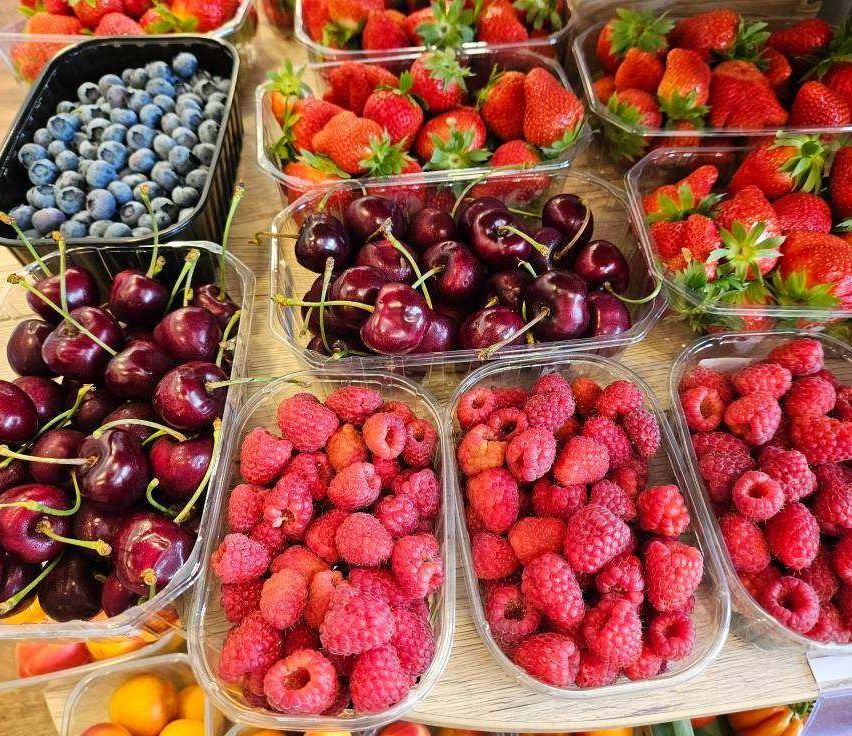 Verschiedene Beeren wie Erdbeeren, Kirschen, Blaubeeren und Himbeeren sind in Plastikbehältern auf einer Holzoberfläche ausgestellt.