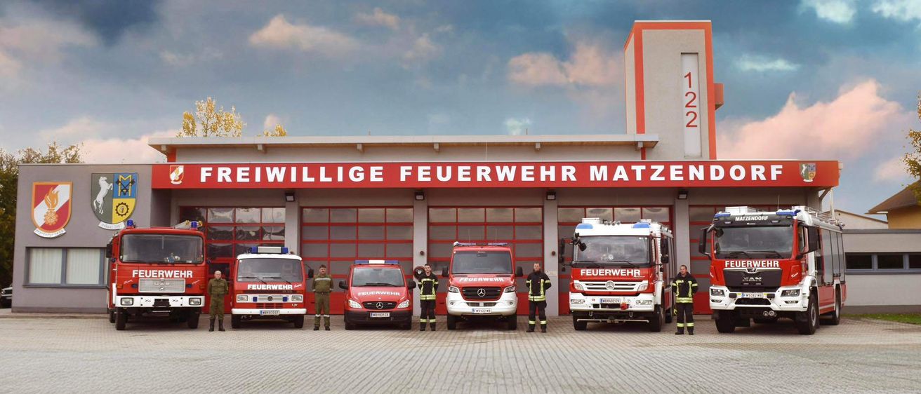 Eine Gruppe Feuerwehrleute steht vor einer Feuerwache, mehrere Feuerwehrfahrzeuge sind geparkt. Das Schild sagt 'Freiwillige Feuerwehr Matzendorf'.