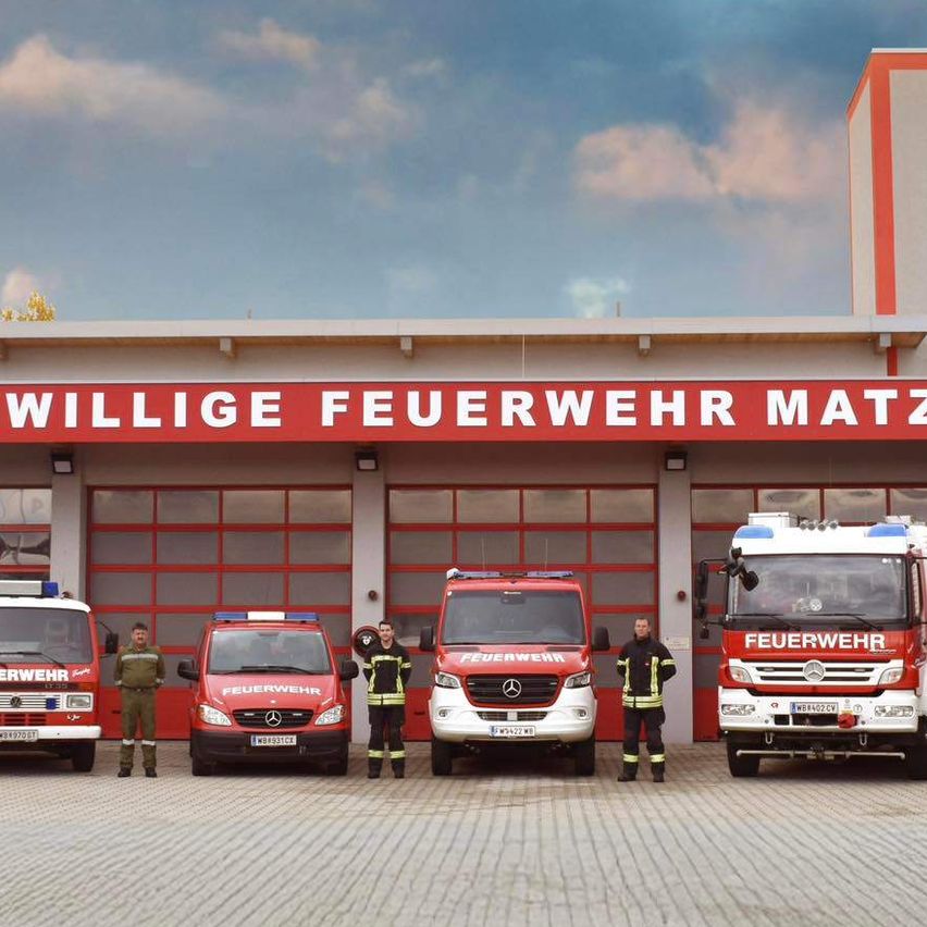 Eine Gruppe Feuerwehrleute steht vor einer Feuerwache, mehrere Feuerwehrfahrzeuge sind geparkt. Das Schild sagt 'Freiwillige Feuerwehr Matzendorf'.