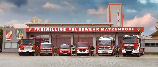 Eine Gruppe Feuerwehrleute steht vor einer Feuerwache, mehrere Feuerwehrfahrzeuge sind geparkt. Das Schild sagt 'Freiwillige Feuerwehr Matzendorf'.