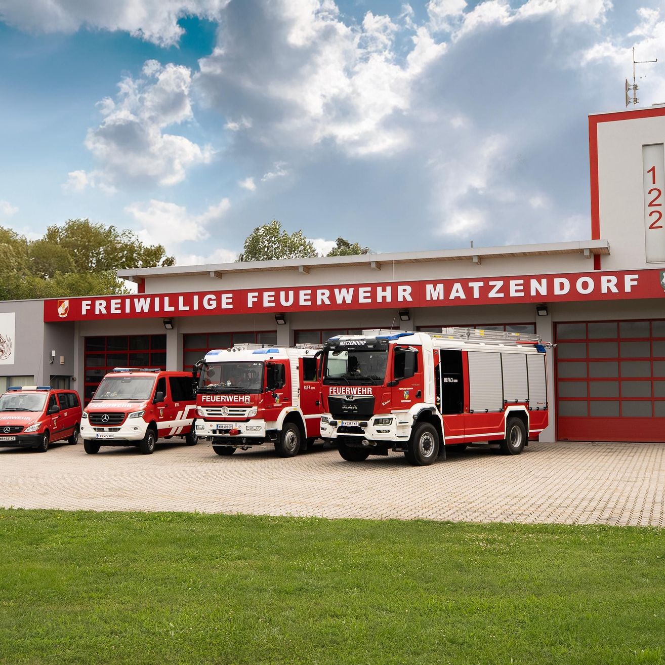 Feuerwehrfahrzeuge stehen vor der Freiwilligen Feuerwehr Matzendorf. Die Fahrzeuge sind rot und weiß, eines hat 'Feuerwehr' darauf geschrieben.