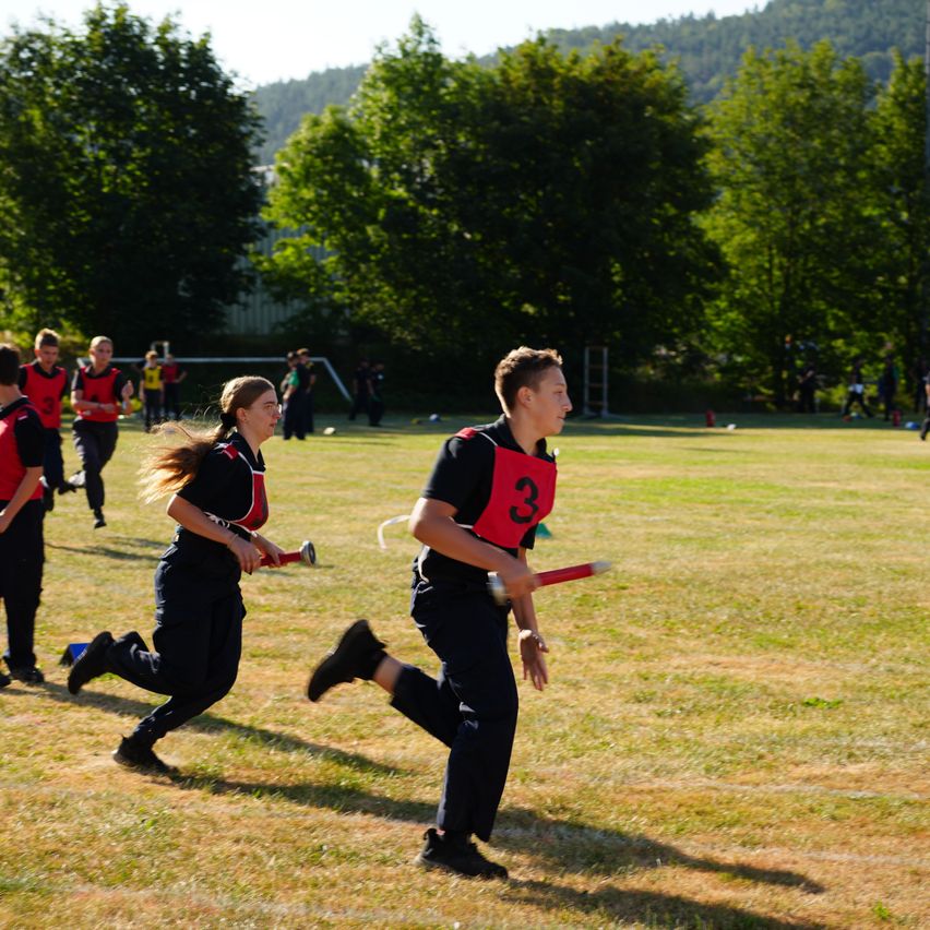 Eine Gruppe junger Athleten läuft auf einem Grasfeld und trägt rote und schwarze Uniformen. Sie halten rote Stöcke, wahrscheinlich für ein Spiel oder Training. Im Hintergrund sind Bäume und ein Gebäude zu sehen.