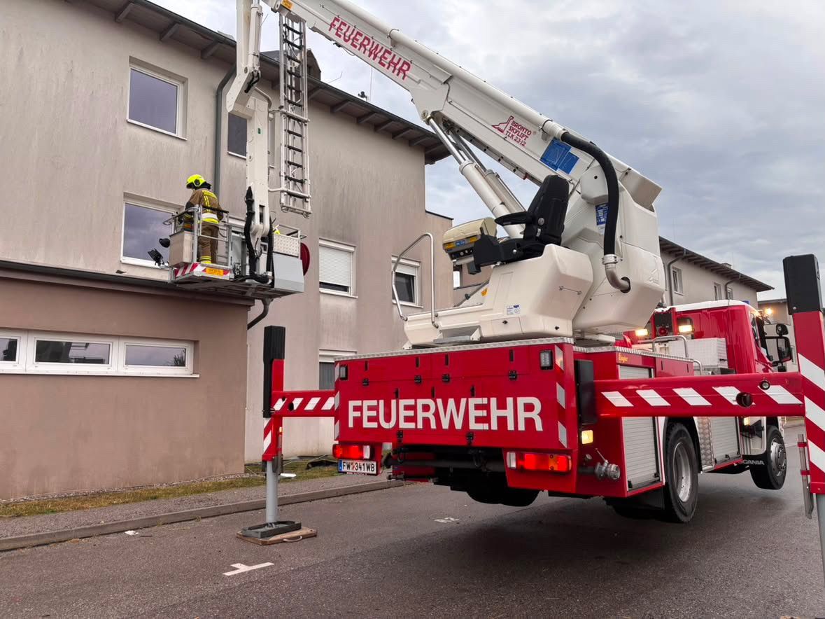 Ein Feuerwehrmann benutzt einen weißen und roten Kran, um ein Fenster im zweiten Stock eines Gebäudes zu erreichen. Der Kran ist mit 'Feuerwehr' beschriftet.