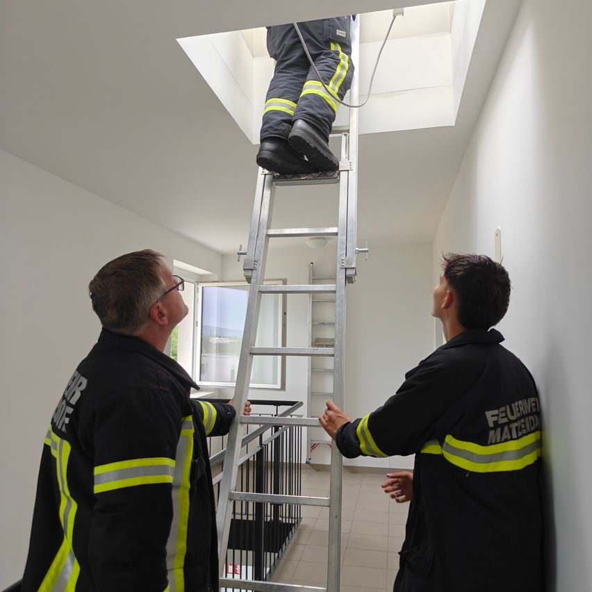 Ein Feuerwehrmann klettert eine Leiter in einem Gebäude hoch, während zwei andere Feuerwehrleute von unten zusehen.