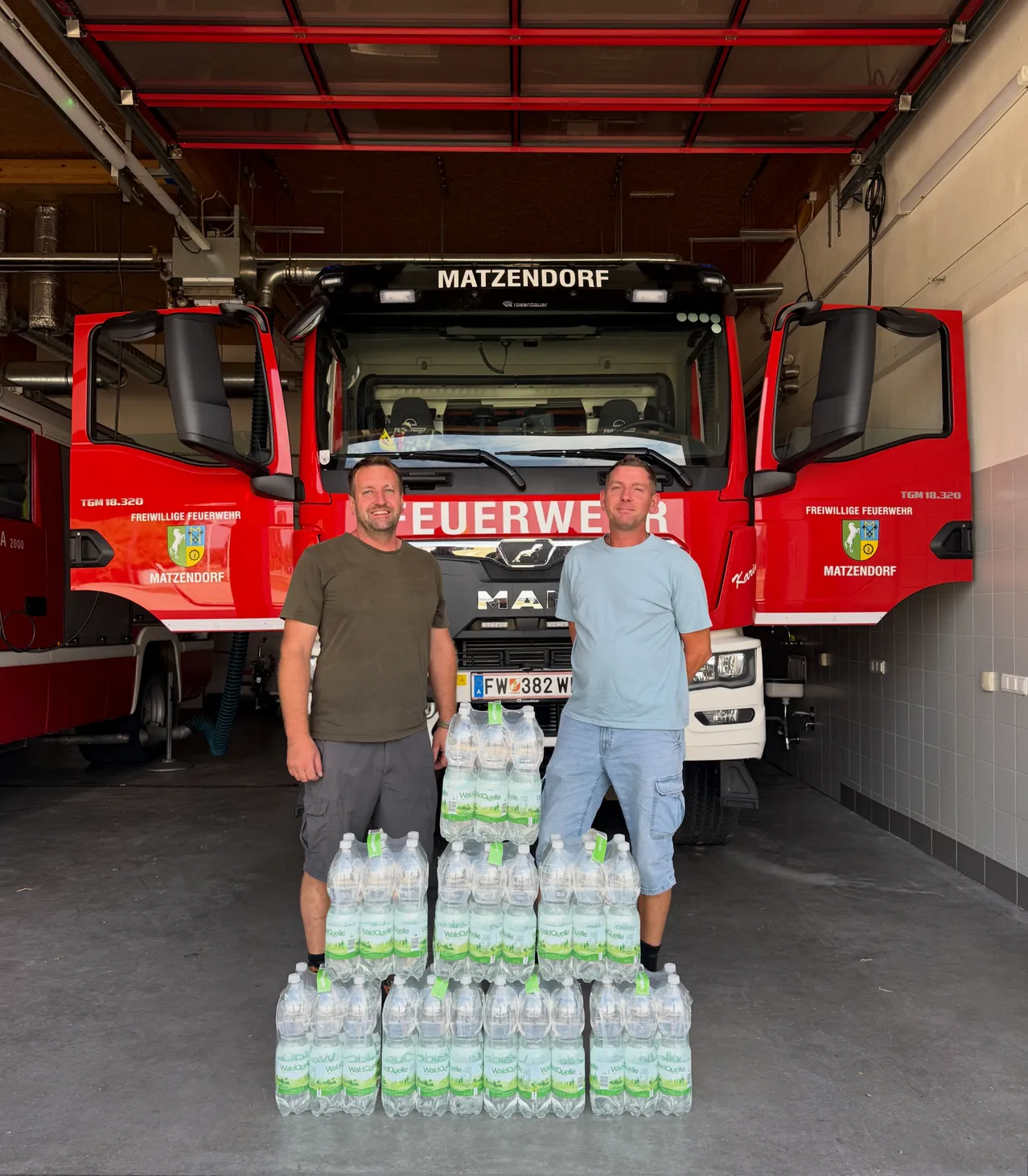 Zwei Männer stehen vor einem Feuerwehrauto mit einem großen Stapel Wasserflaschen. Sie lächeln und scheinen für ein Foto zu posieren. Das Feuerwehrauto ist mit 'MATZENDORF' beschriftet.