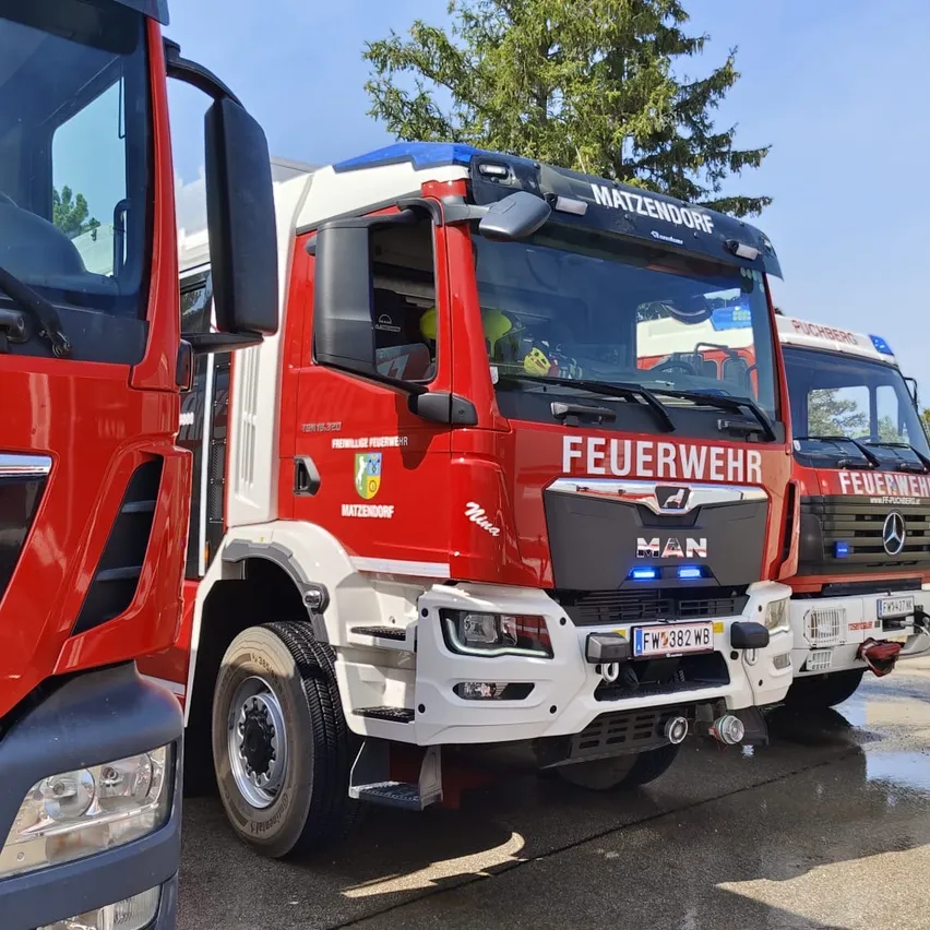 Drei rote Feuerwehrfahrzeuge mit der Aufschrift 'Feuerwehr' sind in einer Reihe auf einer nassen Straße geparkt. Das mittlere Fahrzeug hat 'MATZENDORF' auf der Seite.