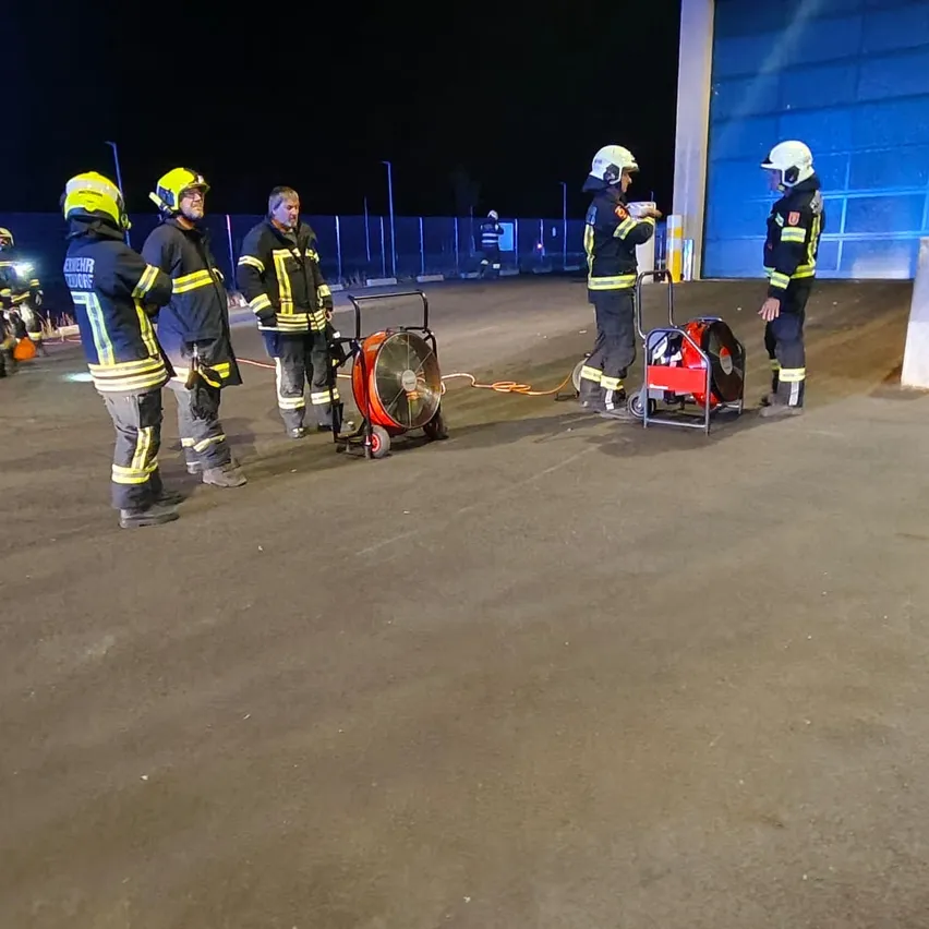 Fünf Feuerwehrleute in voller Ausrüstung stehen nachts draußen, zwei mit Ventilatoren und einer hält ein Papier.