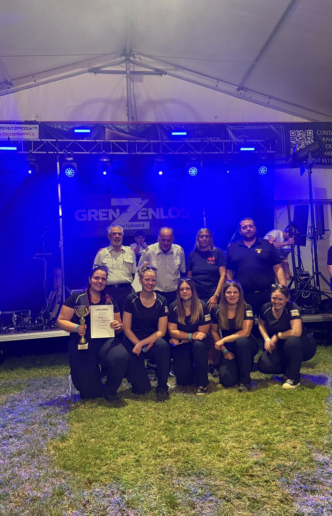 Eine Gruppe von Menschen, darunter Frauen in Uniform, posiert für ein Foto auf einer Bühne mit blauen Lichtern. Eine Frau vorne hält einen Pokal und ein Zertifikat. Hinter ihnen befindet sich ein Banner mit dem Text 'GRENZENLOS'.