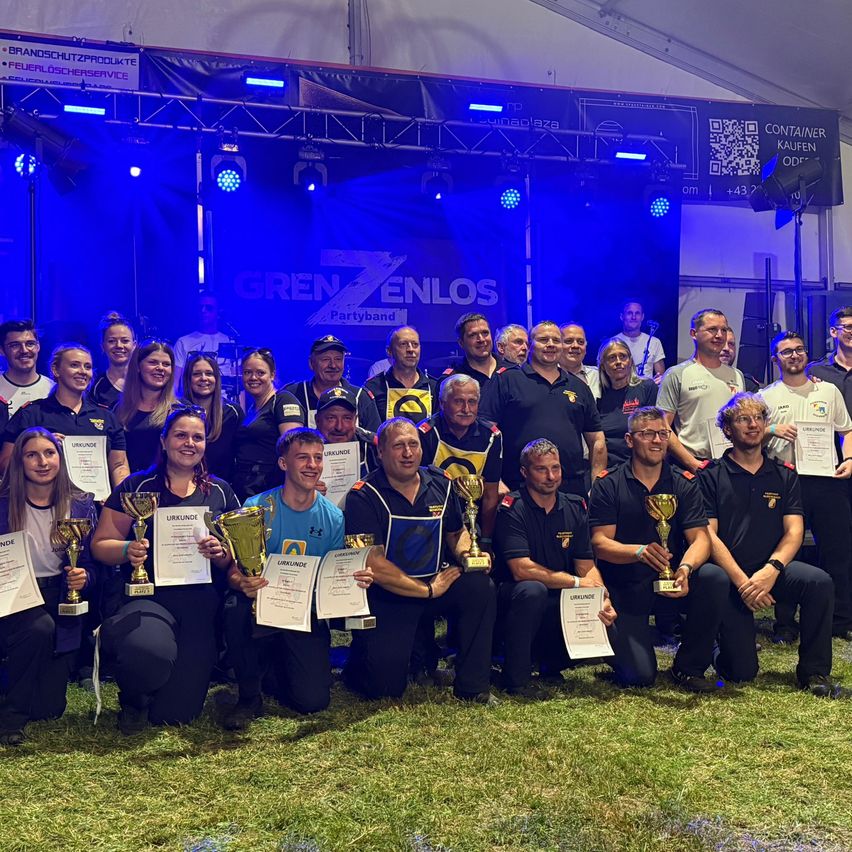 Eine Gruppe von Menschen in passenden Uniformen posiert mit Trophäen und Zertifikaten vor einer Bühne mit blauem Hintergrundlicht und einem Banner, das 'FrenZ Enlos Partyband' besagt.