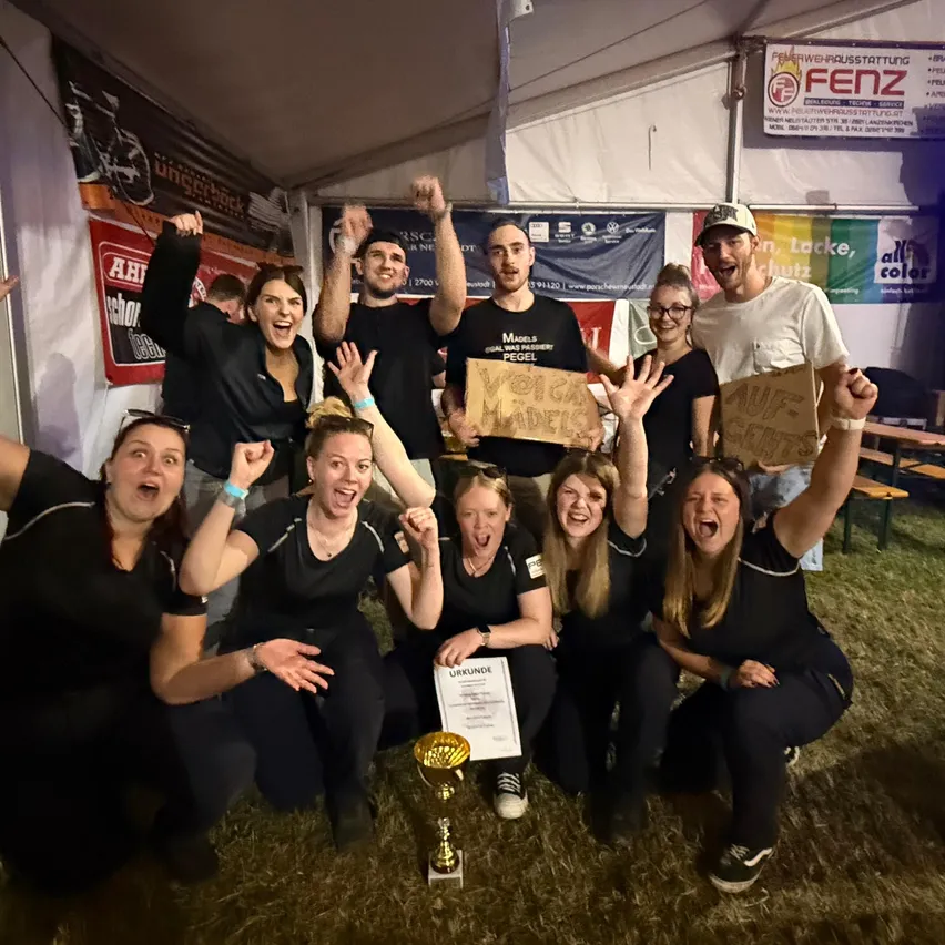 Eine Gruppe von Menschen in schwarzen Uniformen posiert für ein Foto mit einem Pokal und Schildern, in einem Zelt mit Bannern und einer weißen Decke.