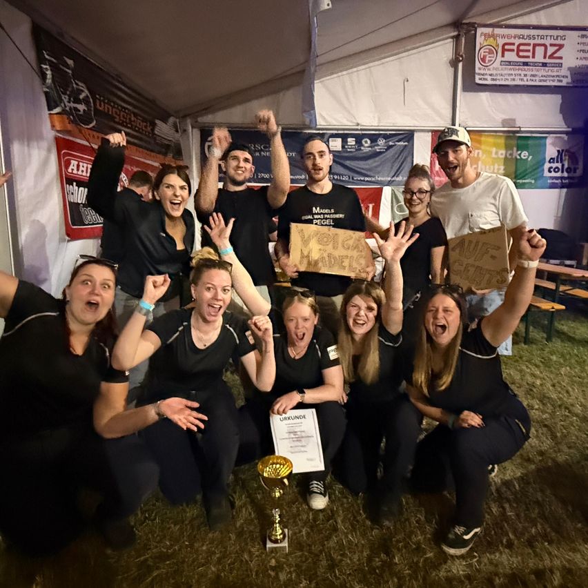 Eine Gruppe von Menschen in schwarzen Uniformen posiert für ein Foto mit einem Pokal und Schildern, in einem Zelt mit Bannern und einer weißen Decke.