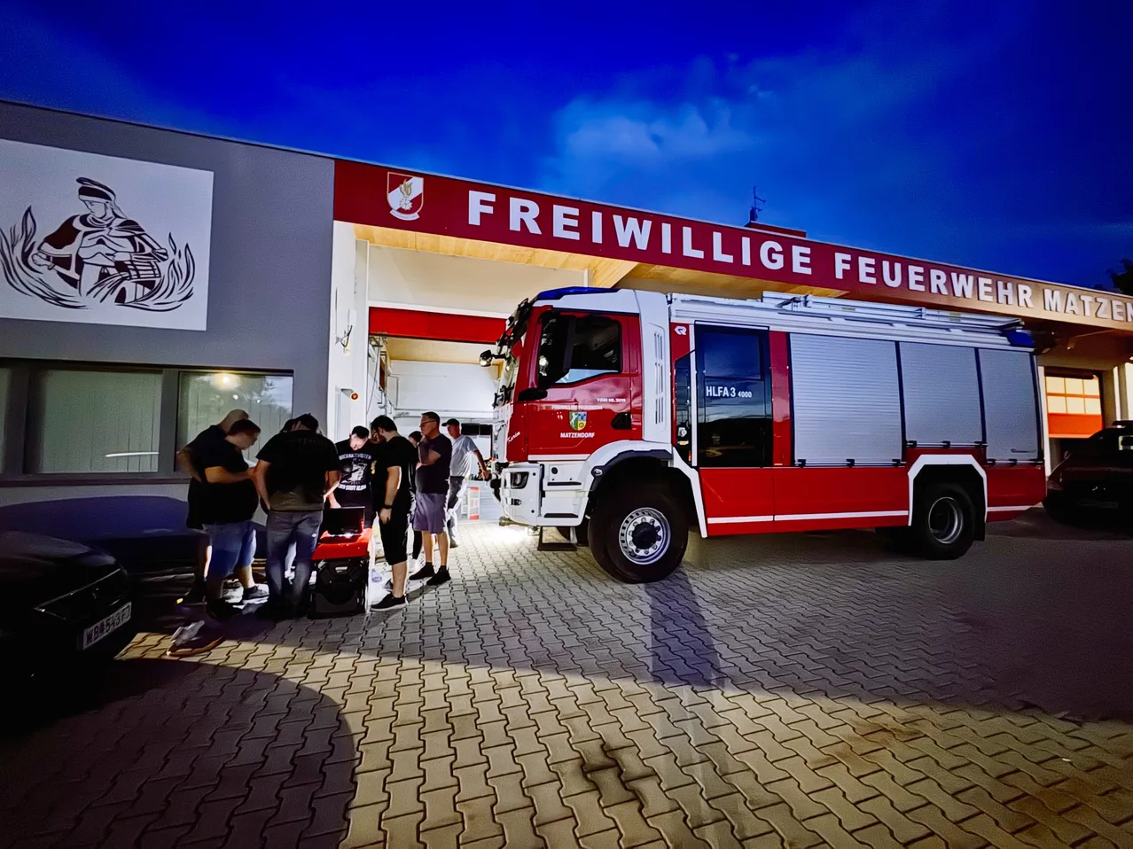Eine Gruppe von Menschen versammelt sich um einen Feuerwehrwagen, der nachts vor einem Feuerwehrhaus geparkt ist.