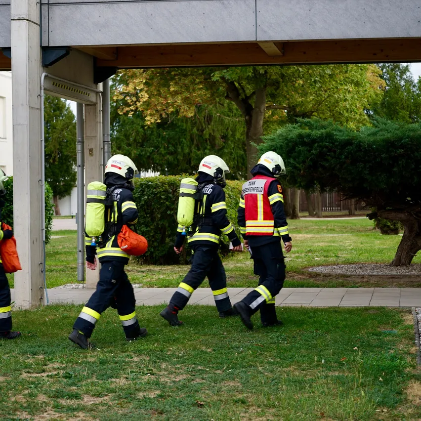 Drei Feuerwehrleute in gelben und roten Uniformen gehen auf einem Rasen, möglicherweise in einem Park. Sie tragen Helme und tragen Rucksäcke.