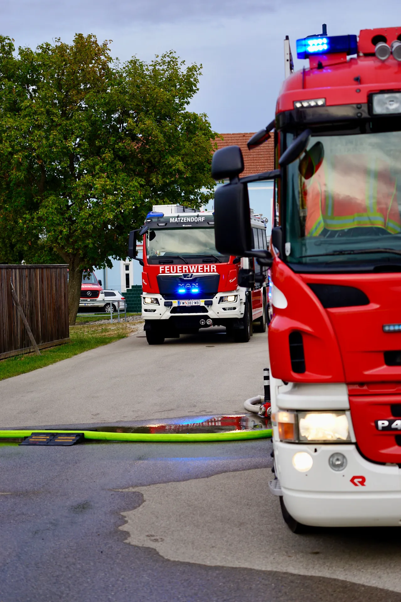 Ein roter Feuerwehrwagen mit blauen Lichtern steht am Straßenrand. Ein grüner Schlauch liegt auf der Straße. Ein Baum befindet sich auf der linken Seite.