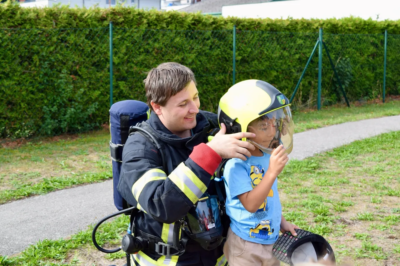 Ein Feuerwehrmann hilft einem kleinen Jungen, einen gelben Helm aufzusetzen. Der Junge trägt ein blaues Shirt mit Comicfiguren. Sie stehen auf einem Rasen.