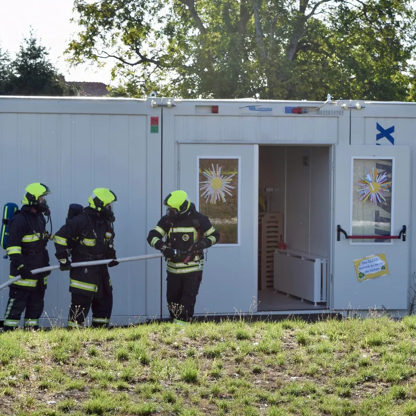 Drei Feuerwehrleute in voller Ausrüstung stehen vor einem weißen Container mit offener Tür und halten einen Schlauch. Der Container hat ein blaues X auf der Seite und ein Fenster mit einem Sonnendesign.