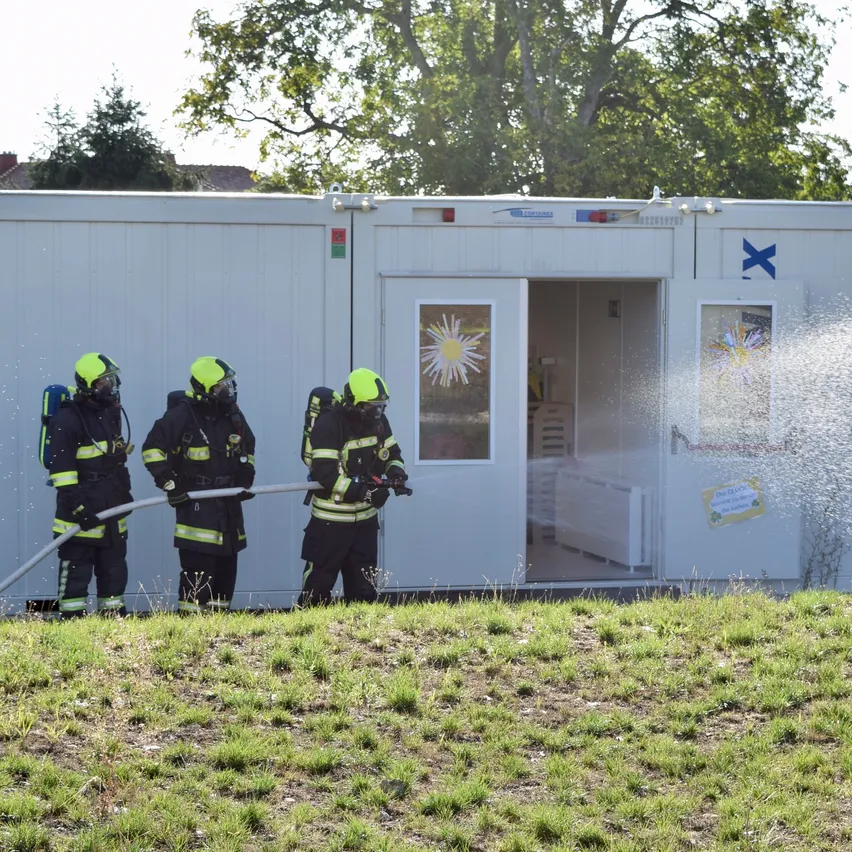 Drei Feuerwehrleute in voller Ausrüstung spritzen Wasser aus einem Schlauch auf ein weißes Gebäude mit offener Tür.