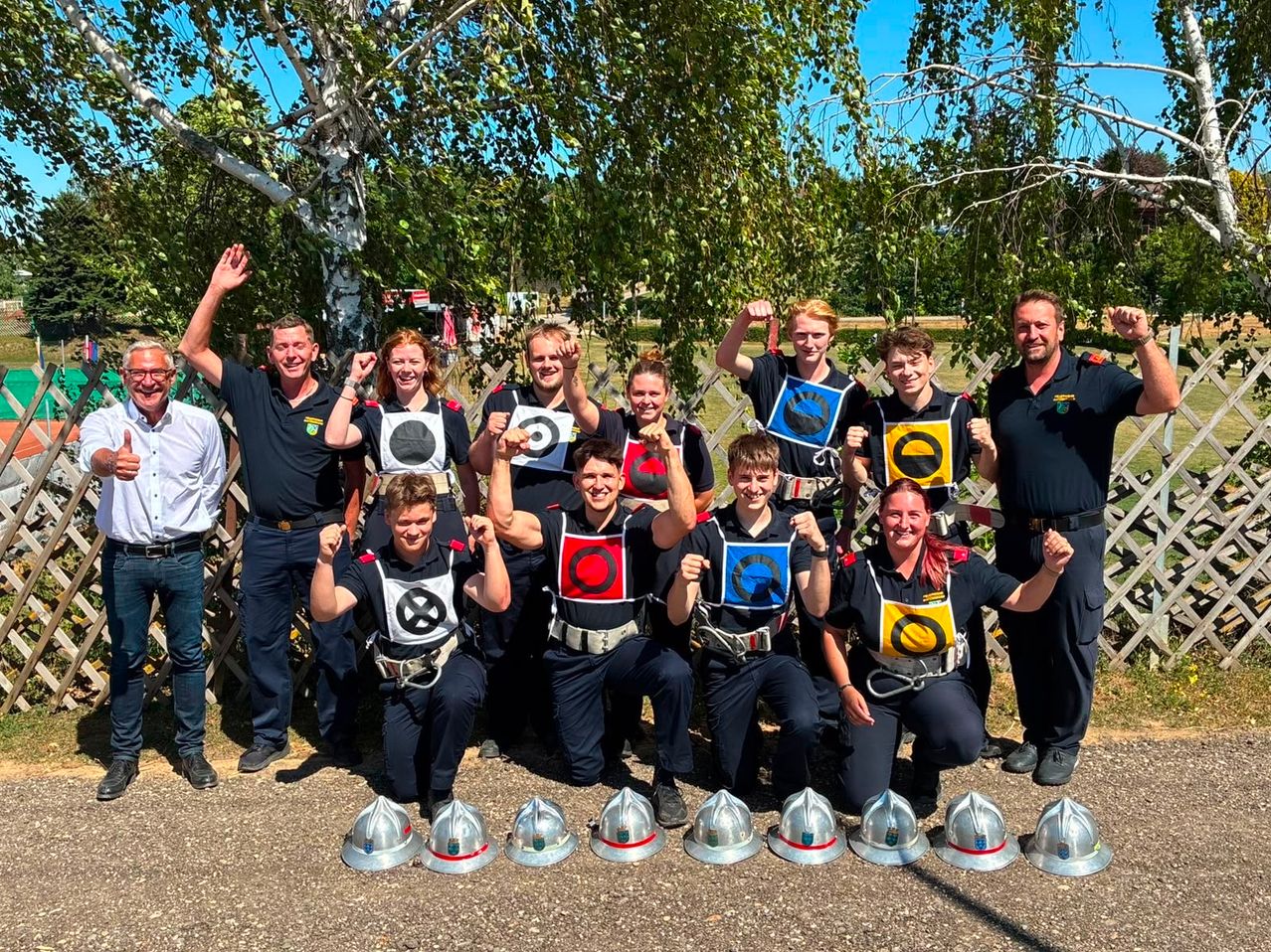 Eine Gruppe von Menschen in Uniformen mit Abzeichen und Helmen posiert für ein Foto im Freien. Sie stehen und knien und halten Schilder mit Designs. Sie lächeln und heben die Hände. Hinter ihnen stehen Bäume, und in der Ferne sind Gebäude und ein klarer Himmel zu sehen.