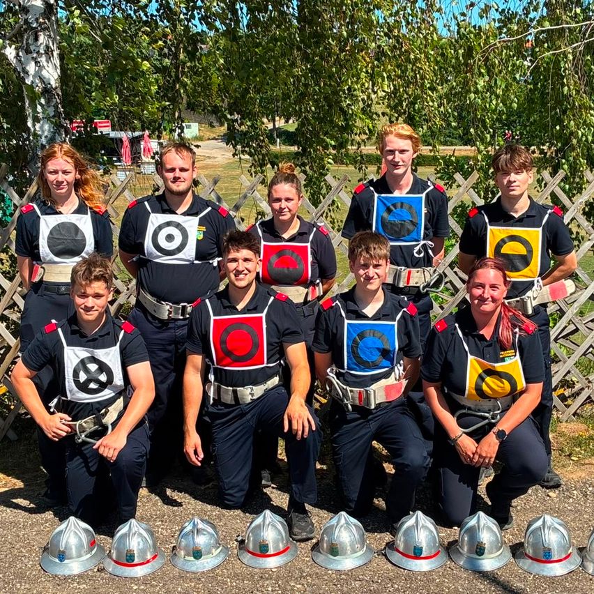 Eine Gruppe junger Feuerwehrleute in Uniform, die lächelnd für ein Foto posiert und mit Helmen vor ihnen in einem Park mit Bäumen und einem Zaun.